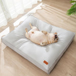 A light gray square dog bed on a light wood floor, with a light-colored French bulldog lying on its back inside the bed. A small orange tag is visible in the bottom right corner of the bed.