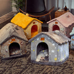 Four plush house-shaped pet beds in different colors and patterns are arranged on a patterned rug in a living room. One small white dog is lying inside the gray star-patterned pet bed in the foreground.